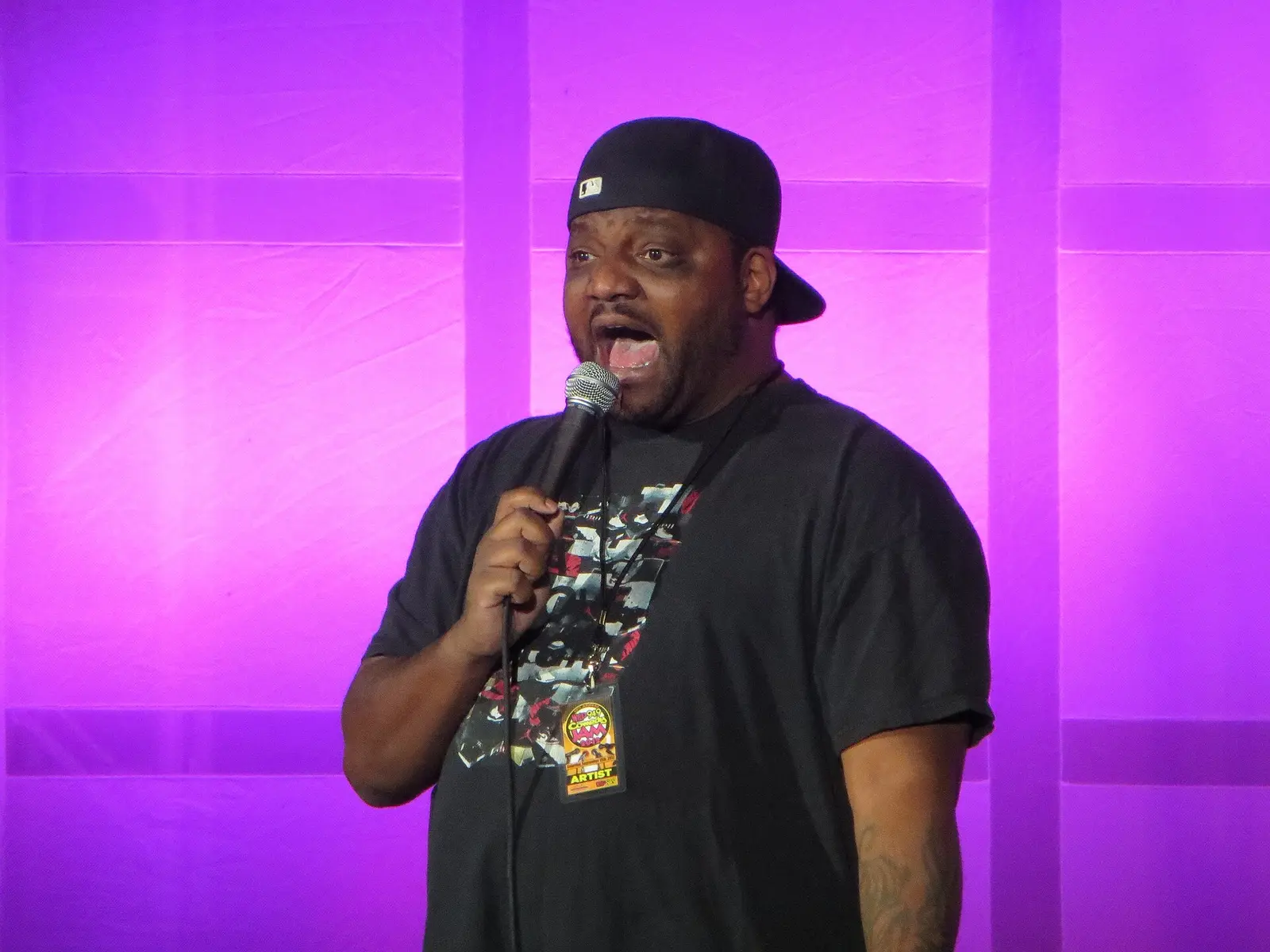Aries Spears performing stand-up in a cap with a microphone against a purple backdrop