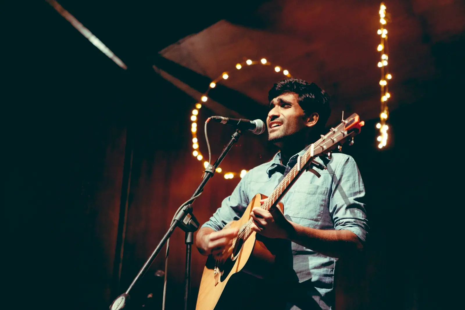 Prateek Kuhad singing and playing an acoustic guitar on a dark stage hung with string lights