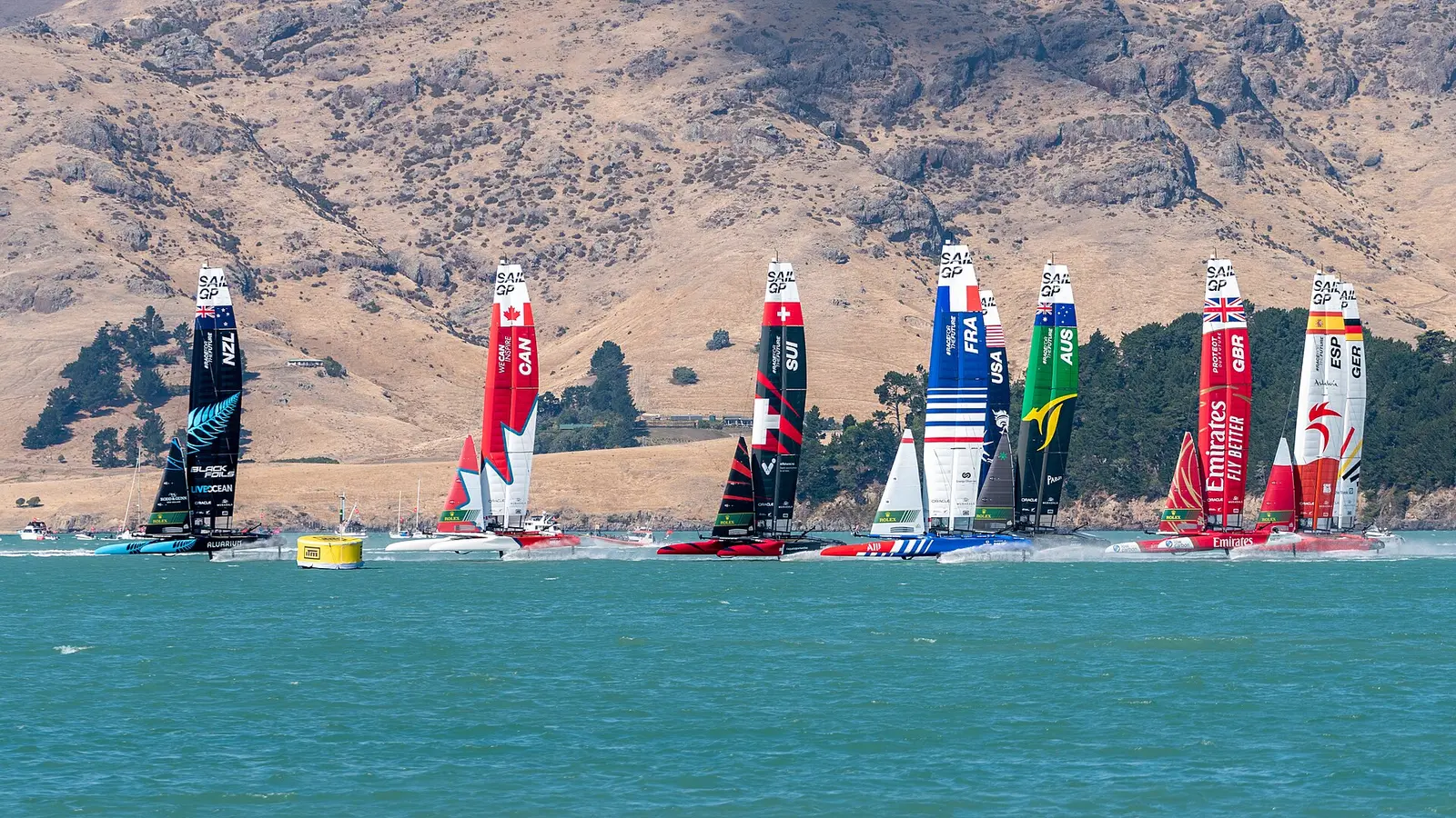 A fleet of SailGP foiling catamarans racing off a hilly coast