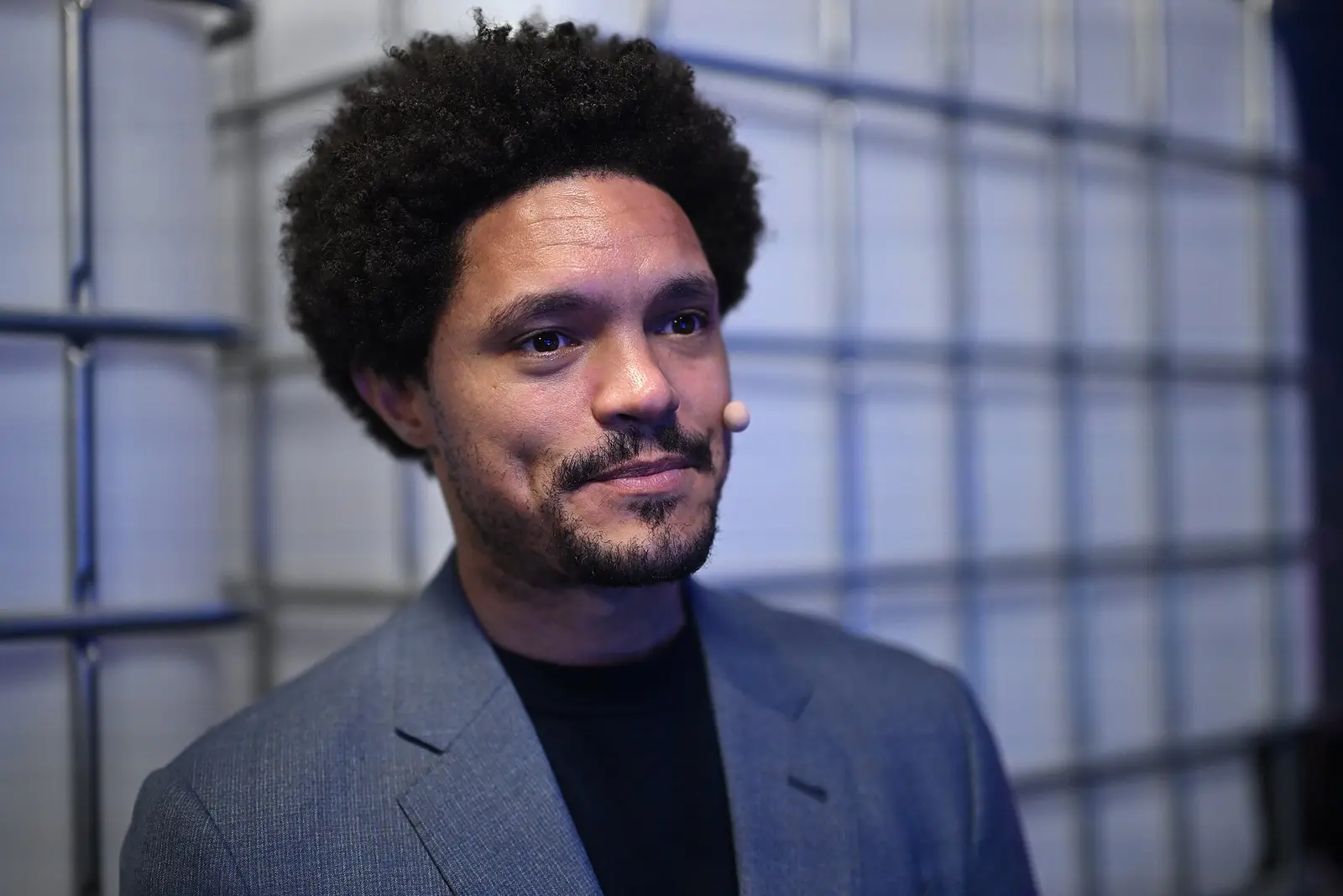 Trevor Noah in a grey blazer wearing a headset microphone