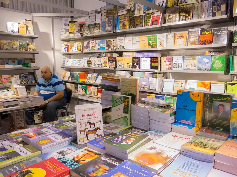 A bookseller seated among stacked books on a stand inside an exhibition hall at Expo Centre Sharjah in 2014